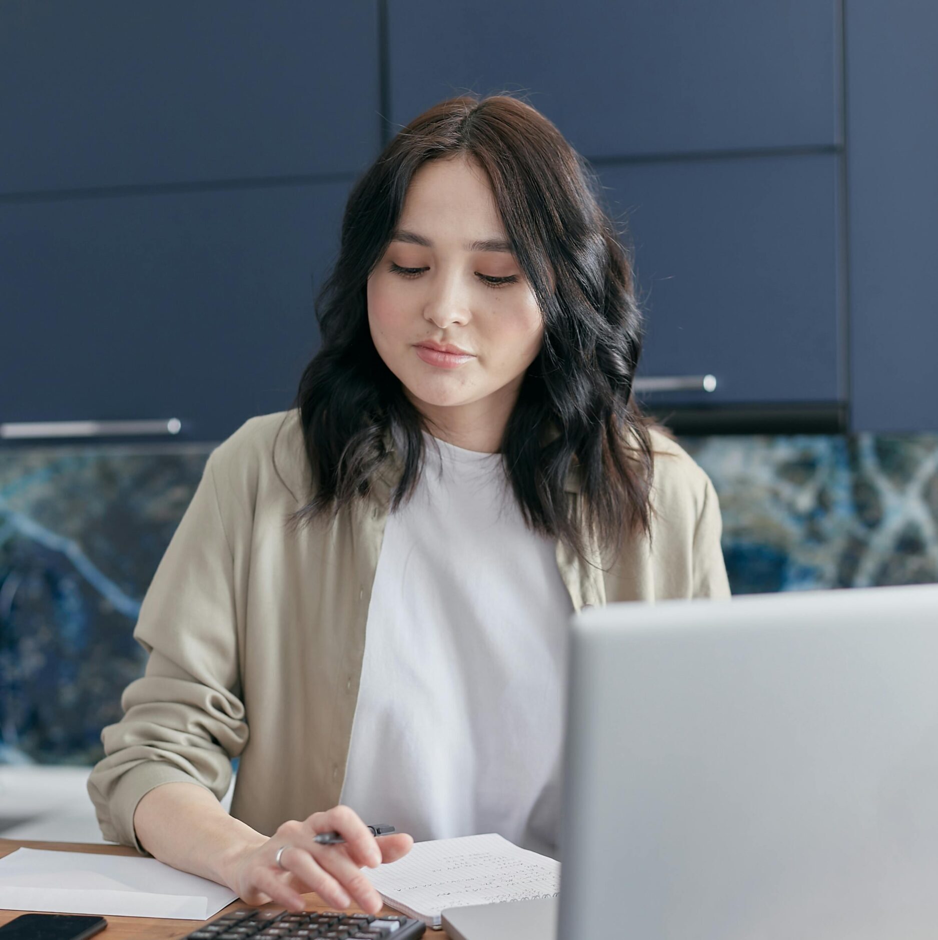 A young woman calculating finances at home using a laptop and calculator, reflecting a focused budgeting process.