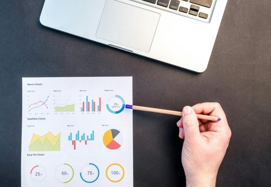 Hand holding pencil reviewing colorful data charts on desk with laptop.